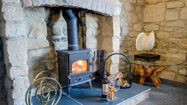 A stone fireplace with a wood-burning stove, a glass table with a bottle and glass, and a wooden stool. - The Anchorage - sea views and beach on doorstep (Amble)