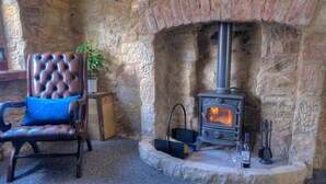 A stone fireplace with a wood-burning stove, a tufted leather armchair, and a small table with a glass. - The Rockpool - Private HOT TUB + Sea Views (Amble)