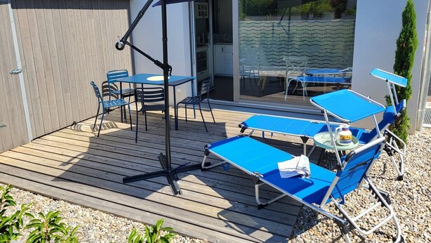 An outdoor patio with a wooden deck, blue lounge chairs, and a table under a blue umbrella.