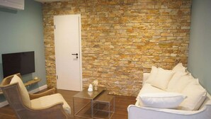 A room with a stone wall, a white door, a white sofa, a wicker chair, a small table, and a flat-screen TV.