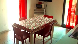 Un comedor con una mesa cubierta por un mantel estampado, rodeada de sillas. Un televisor está montado en la pared y hay un microondas sobre una mesita.