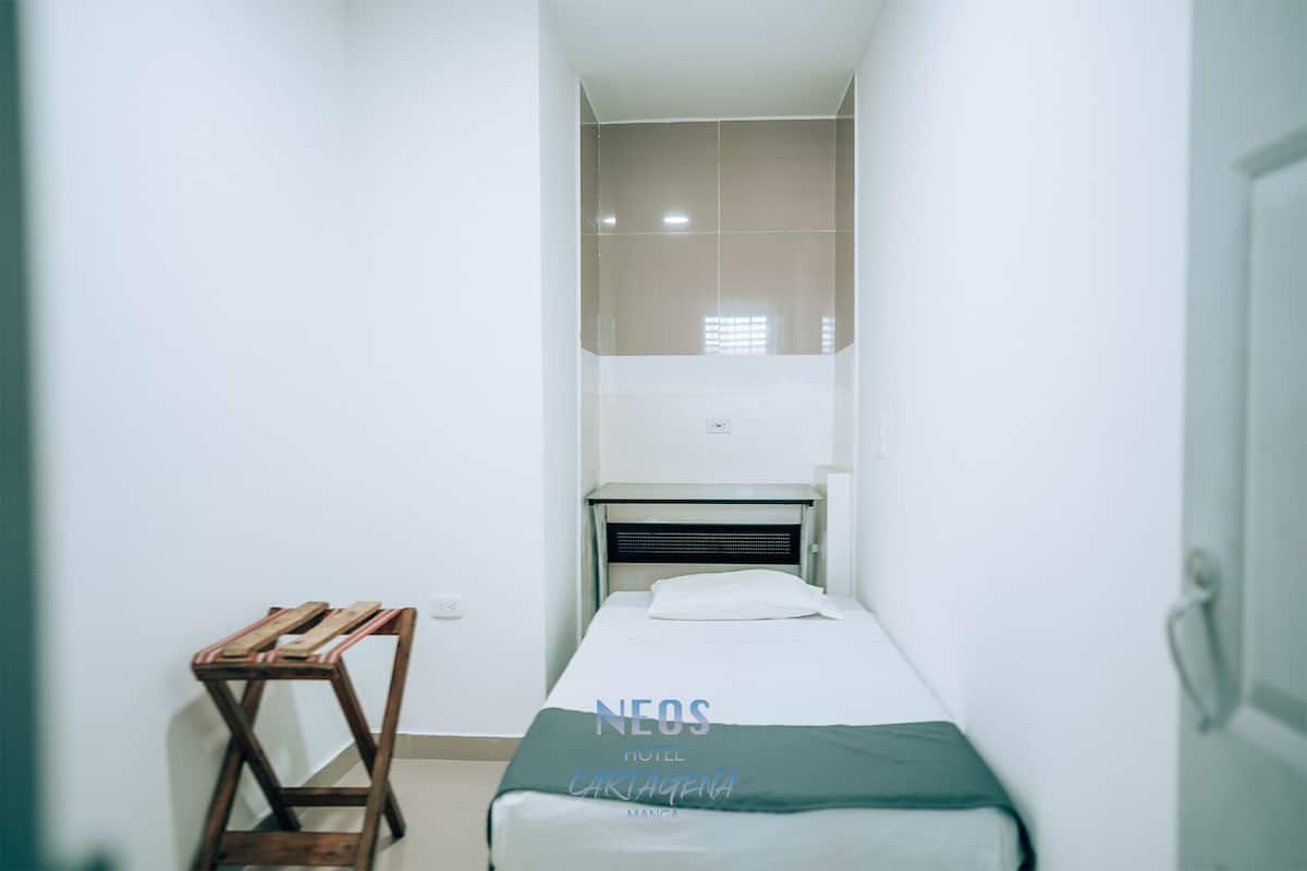 Una habitación de hotel pequeña y estrecha con una cama individual, un taburete de madera y una ventana con persianas.