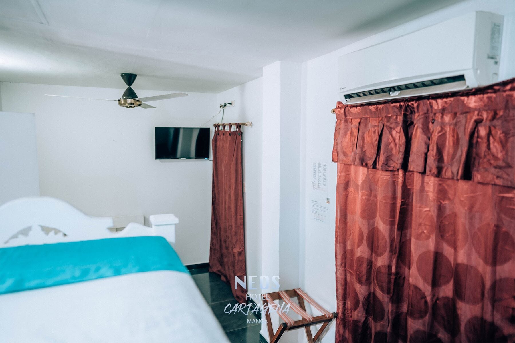 A hotel room with a bed, a TV mounted on the wall, a ceiling fan, and a window with red curtains.