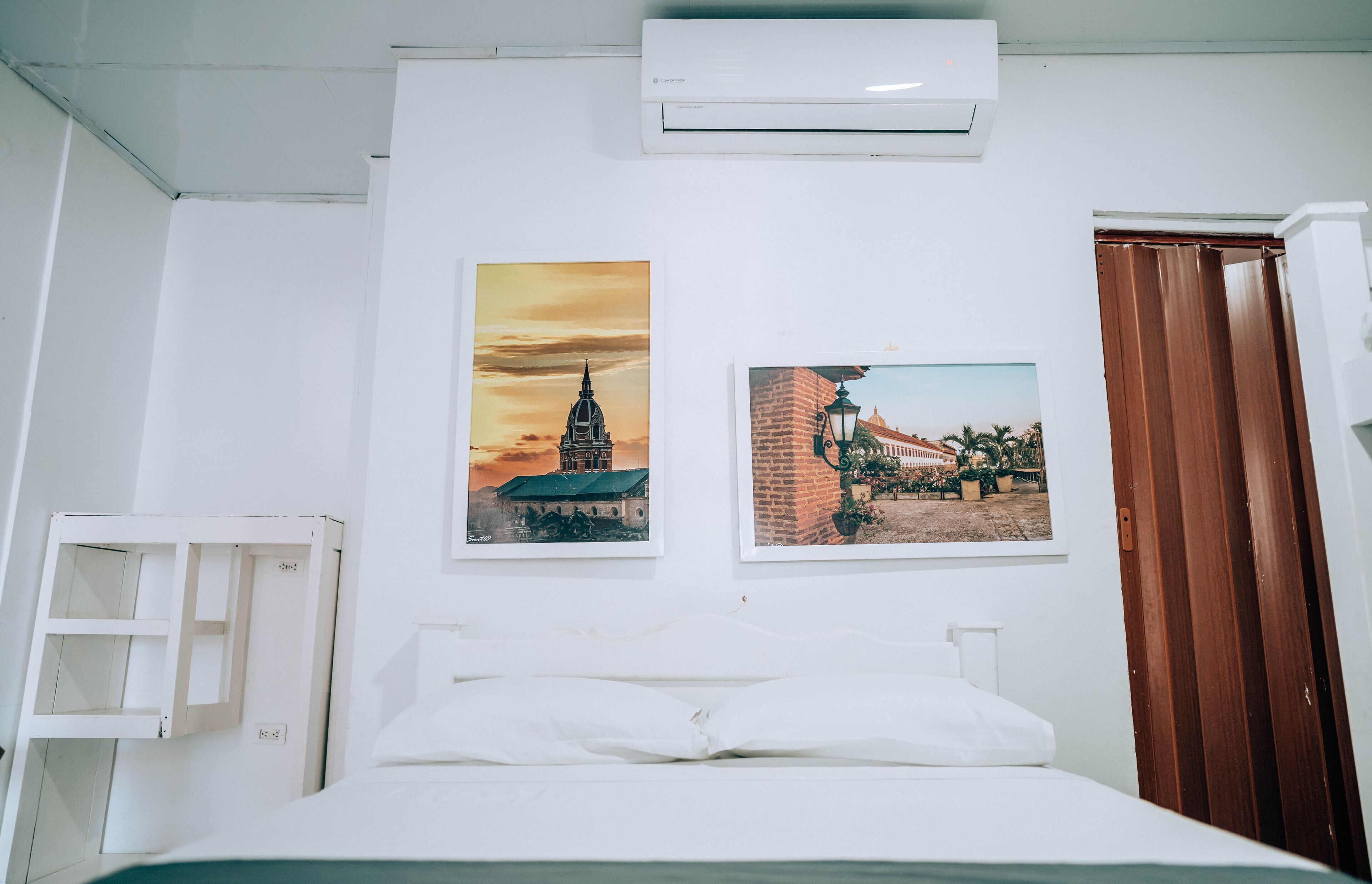 A hotel room with a bed, white walls, two framed pictures, an air conditioner, and a wooden door.