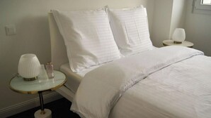 Una cama bien hecha con sábanas blancas, dos mesitas de noche con lámparas y un pequeño florero sobre una de ellas.