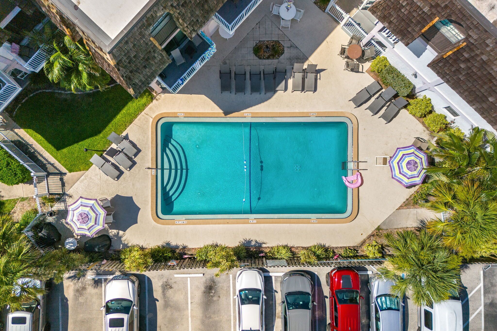 An aerial view of a swimming pool surrounded by lounge chairs, umbrellas, and parked cars.