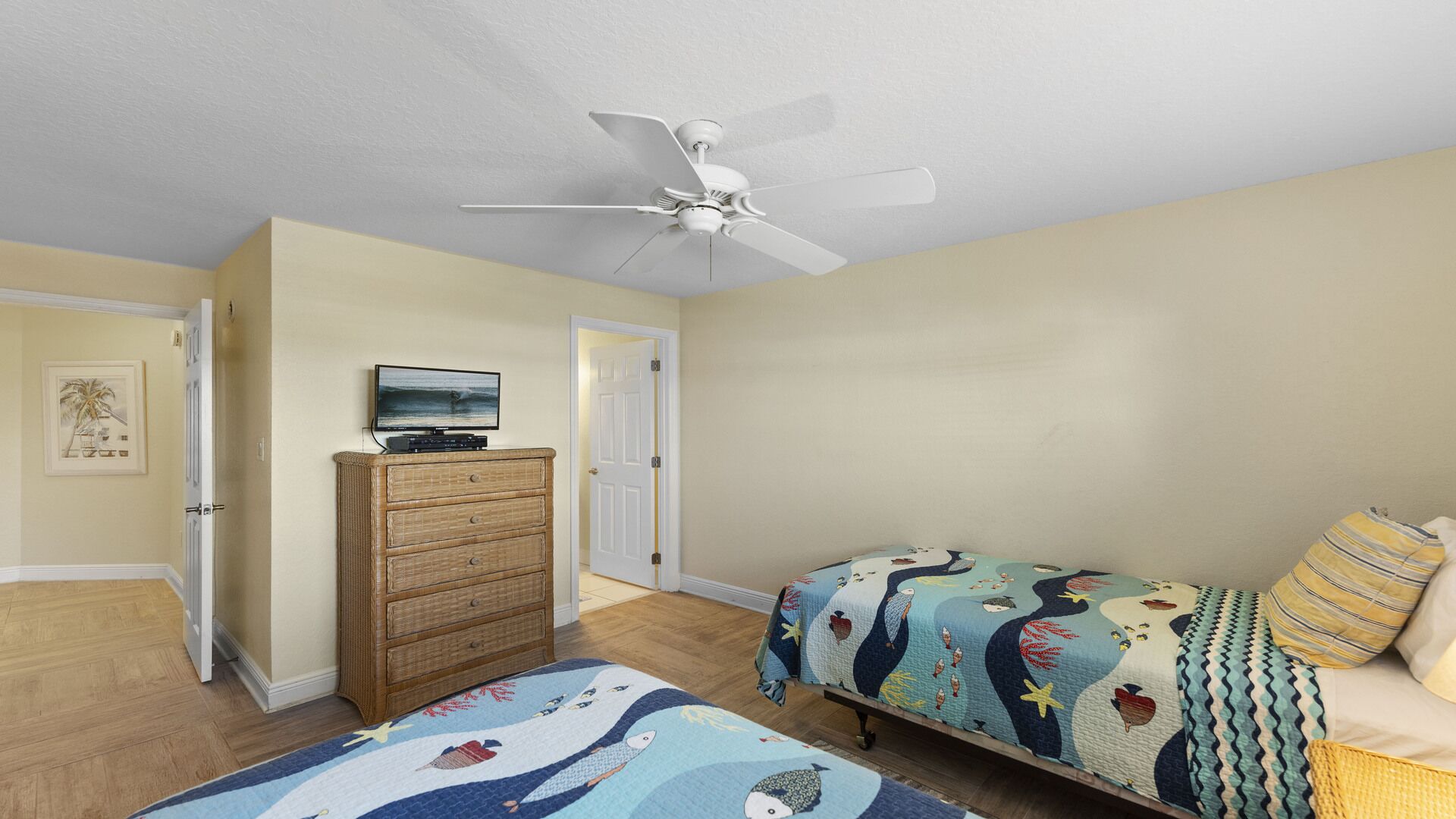 Une chambre à coucher avec un lit, une commode, un téléviseur posé sur la commode, un ventilateur de plafond et une porte.