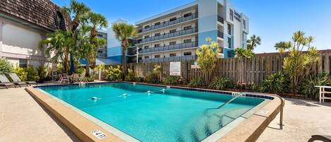 A swimming pool with a wooden deck, surrounded by palm trees and a wooden fence.