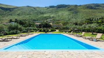 Una piscina rectangular con borde de piedra, rodeada de sillas de descanso y un jardín cuidado con diversas plantas y arbustos.