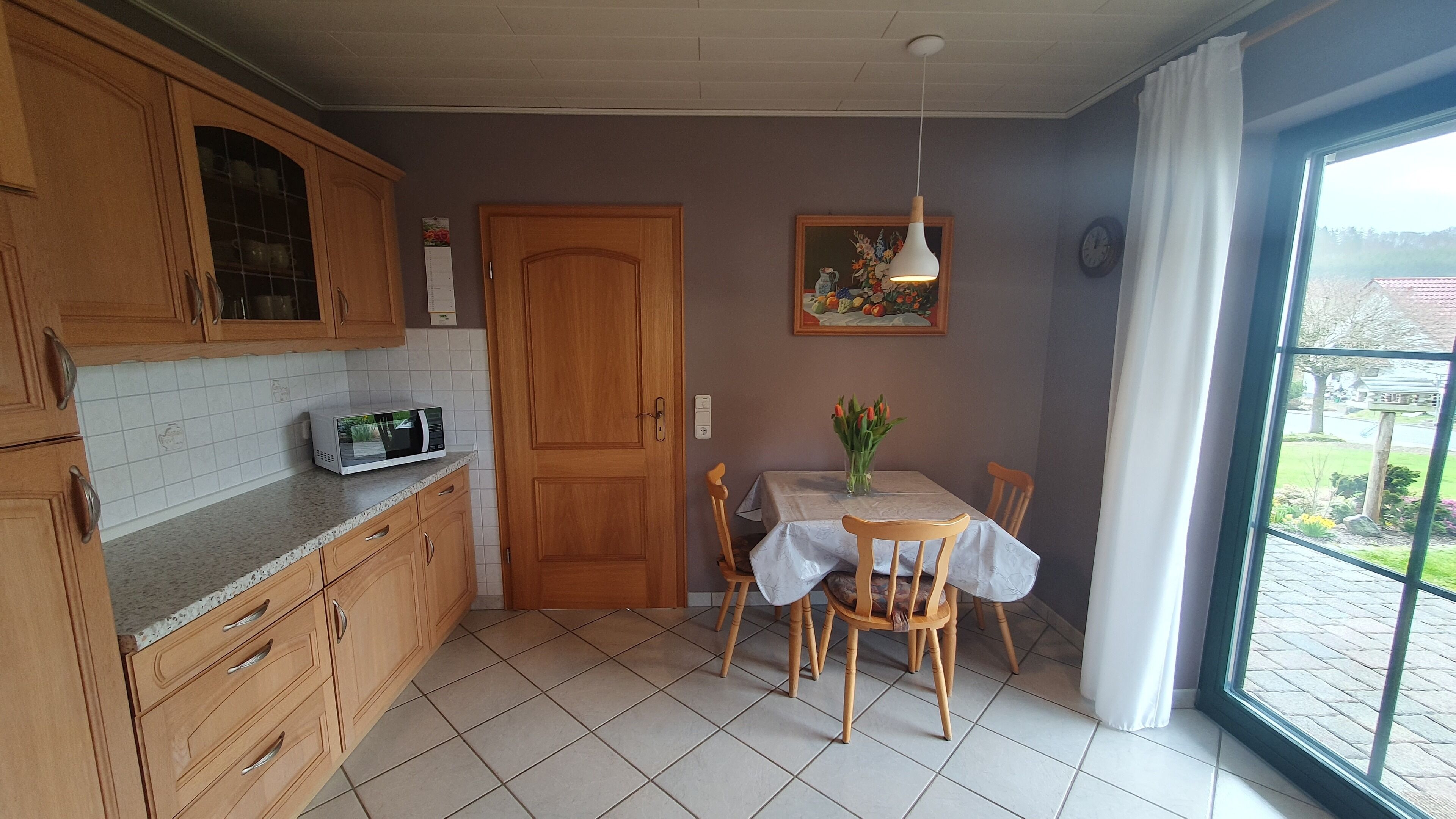 A kitchen with wooden cabinets, a microwave, a dining table with chairs, and a window with a view of the outside.