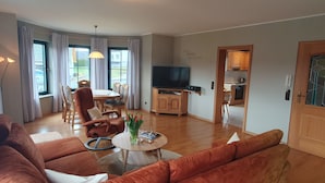 Ein Wohnzimmer mit einer Couch, einem Couchtisch mit Büchern und einer Vase, einem Essbereich mit Tisch und Stühlen, einem Fernseher auf einer Holzkommode und einer durchgehenden Tür erkennbaren Küche.