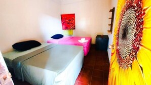 A room with two beds, one with a pink bedspread and the other with a grey one. There is a red abstract painting on the wall, a wooden door with a circular design, and a small table with a white object on it. - Hostal Club Mochilero (La Libertad)