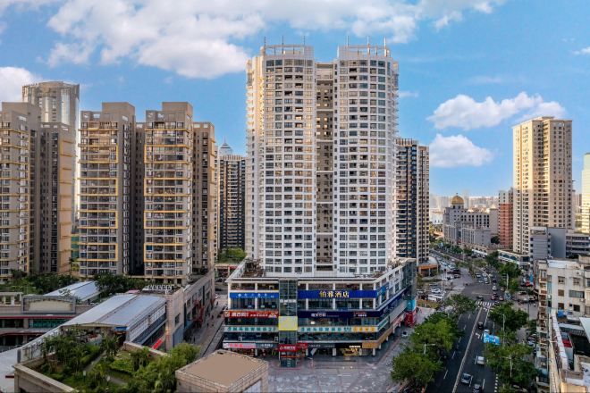 A high-rise residential building with multiple balconies, surrounded by other high-rise buildings, a street with vehicles, and a clear sky.