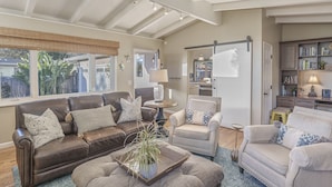 A spacious living room with a brown leather sofa, a grey armchair, and a beige sofa. There is a coffee table with a plant, a lamp on a side table, and a bookshelf with books and decorative items. - 3874 Mission Fields - Adorable Carmel Custom Home (Carmel)