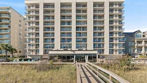 Un hotel de varios pisos con una entrada central, balcones en cada planta y un área de piscina visible al fondo.
