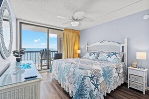 Une chambre avec balcon, un lit recouvert d’une couvre-lit à motifs d’étoiles de mer, un ventilateur de plafond et une table de chevet avec une lampe.