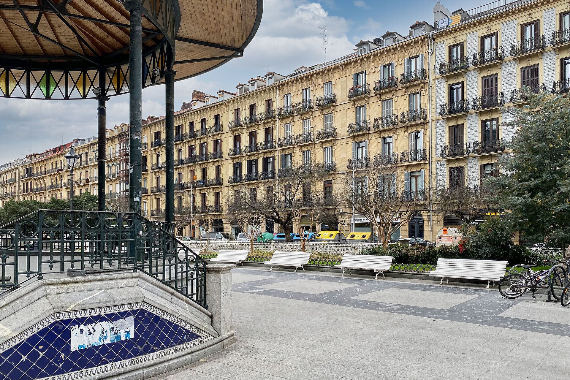 A public square with a tiled fountain, benches, and a row of multi-story buildings with balconies.
