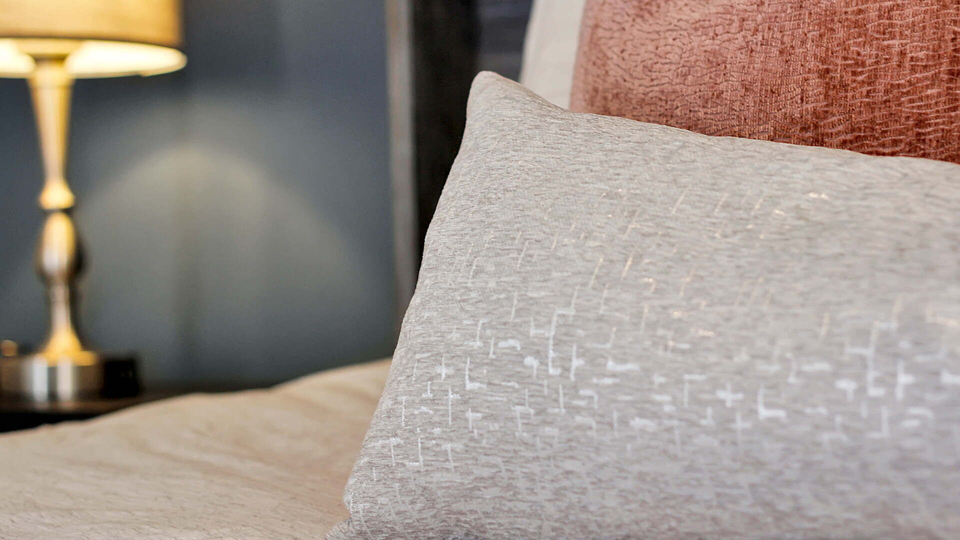 Un gros plan d’un coussin à texture particulière, posé sur un lit, avec une lampe en arrière-plan.