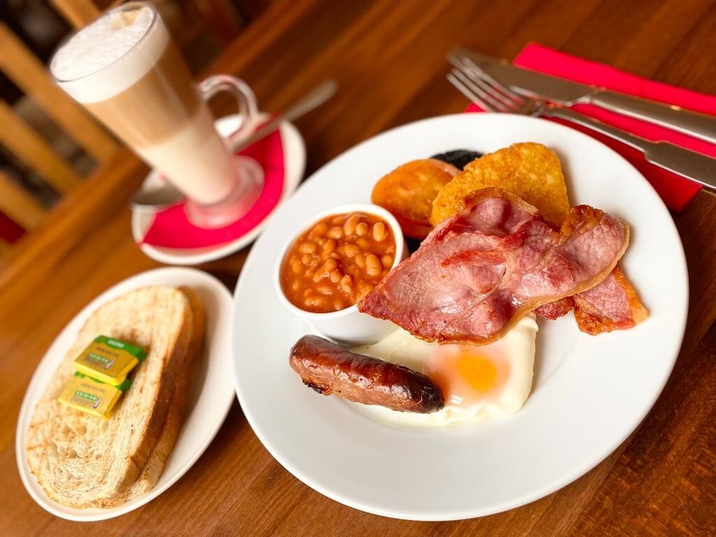 Une assiette de petit-déjeuner comprenant des œufs, du bacon, des saucisses, des haricots, des toasts et une boisson.
