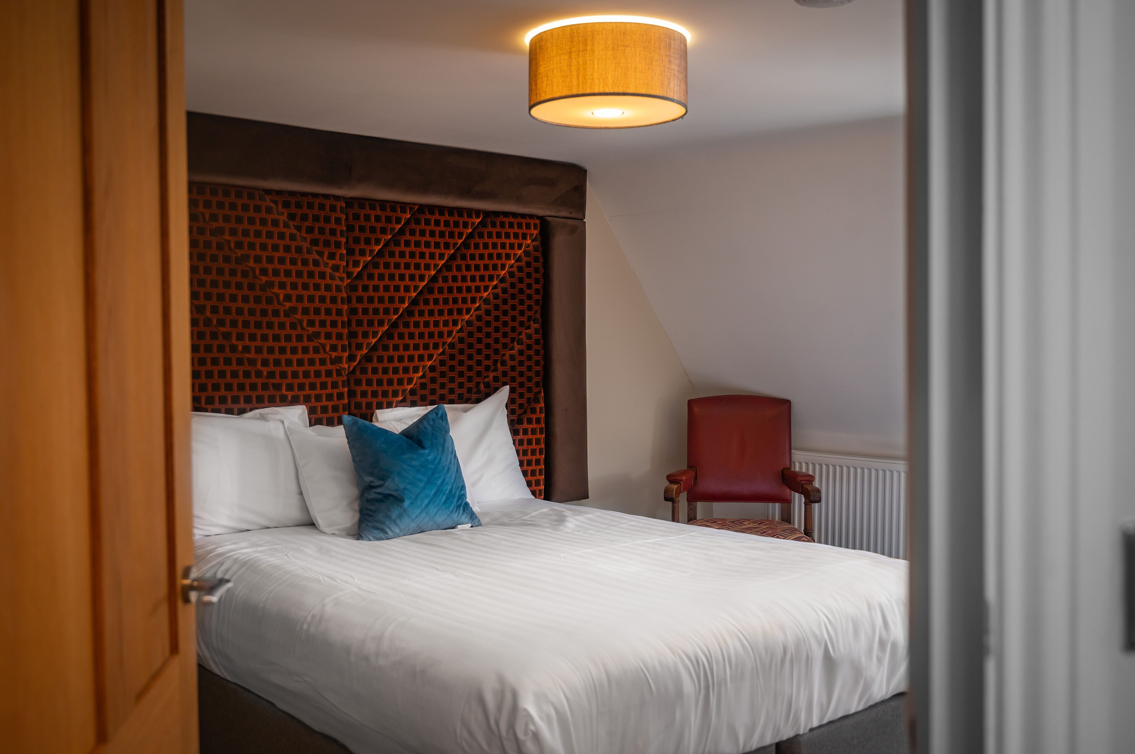 Uma cama bem arrumada com lençóis brancos e almofadas azuis, uma cadeira vermelha e um painel de cama estampado.