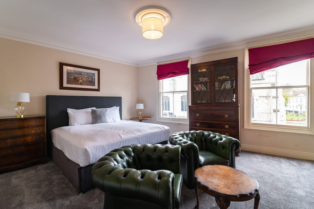 A hotel room with a large bed, two green leather armchairs, a wooden dresser, and a window with red curtains.