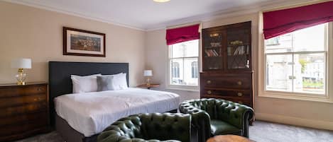 A hotel room with a large bed, two green leather armchairs, a wooden dresser, and a window with red curtains.