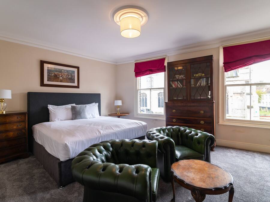 A hotel room with a large bed, two green leather armchairs, a wooden dresser, and a window with red curtains.