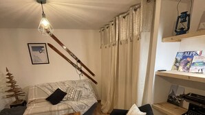 En hyggelig stue med trægulv, en seng med et mønstret tæppe, en sort lænestol, et træ sofabord, en loftslampe, en reol med bøger og et indrammet billede på væggen.