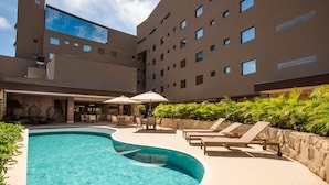 A swimming pool with lounge chairs and umbrellas, surrounded by a building with multiple windows and a stone wall. - Amazon Aeroporto Hotel (Varzea Grande)