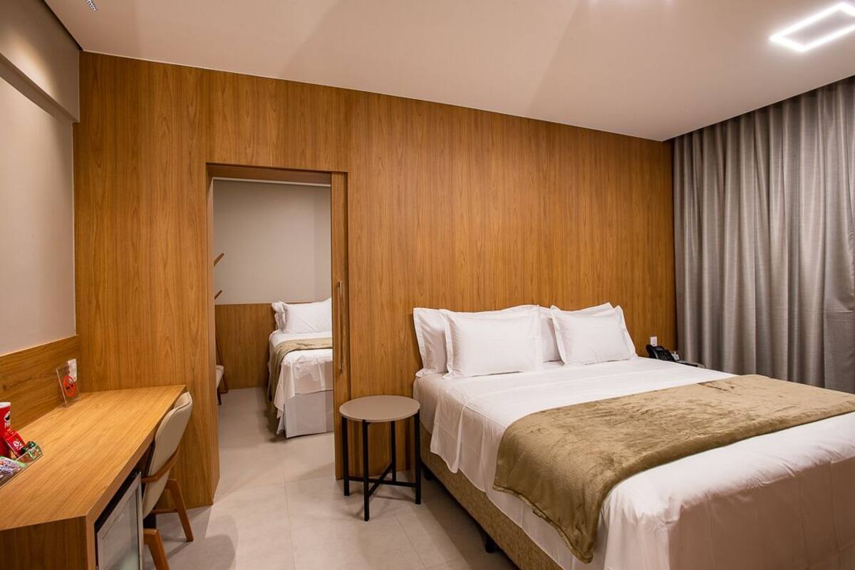 Quarto de hotel com parede de madeira, cama com roupa de cama branca, mesa redonda e escrivaninha.