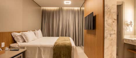 A modern hotel room with a neatly made bed, a flat-screen TV mounted on the wall, a minibar, and a bathroom with a marble wall.