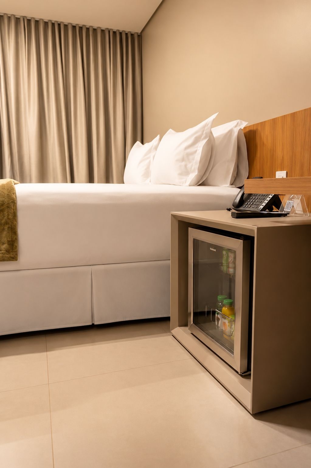 Quarto de hotel com cama, mesa de cabeceira, telefone e mini-geladeira, e cortina.