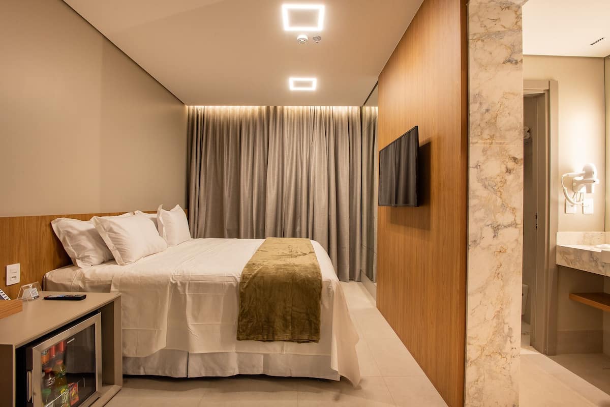 Quarto de hotel com cama, televisão fixada na parede, secretária com computador e casa de banho com espelho e lavatório.