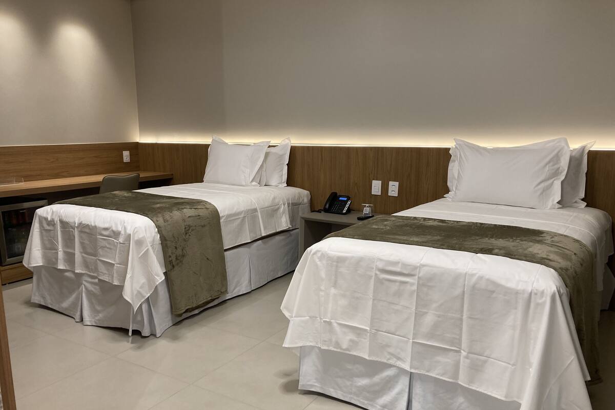 Quarto de hotel com duas camas de solteiro, uma mesa de cabeceira com telefone e uma pequena mesa com abajur.