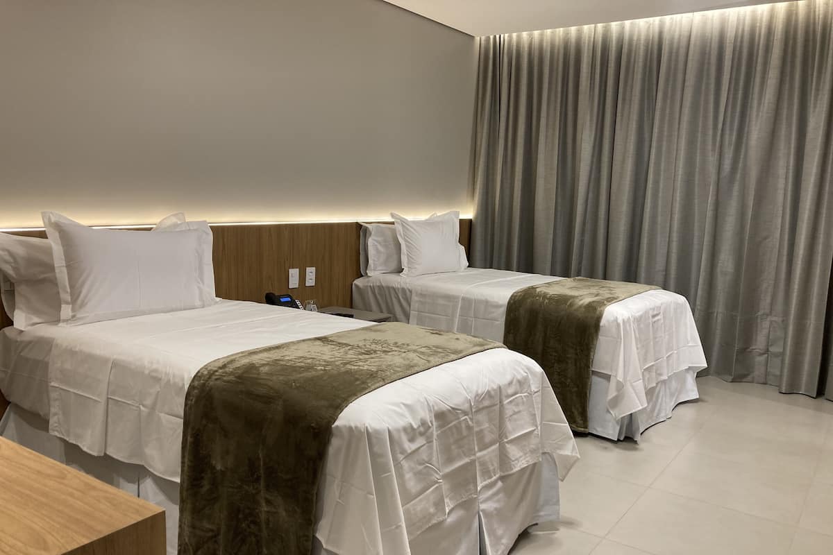 Quarto de hotel com duas camas, uma cabeceira de madeira, mesa de cabeceira e uma janela grande com cortinas.