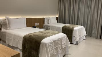 A hotel room with two beds, a wooden headboard, a bedside table, and a large window with curtains.