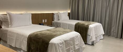 A hotel room with two beds, a wooden headboard, a bedside table, and a large window with curtains.