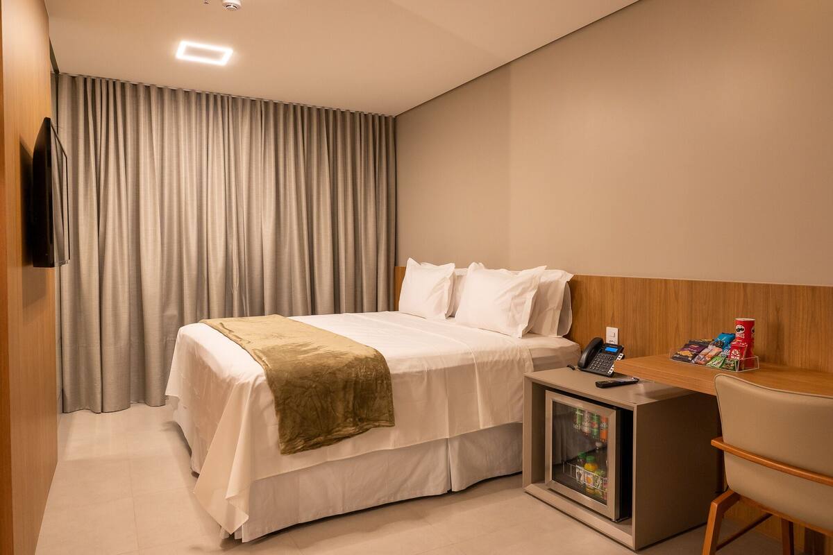 Quarto de hotel com cama, escrivaninha com telefone e lanches, minibar e televisão na parede.