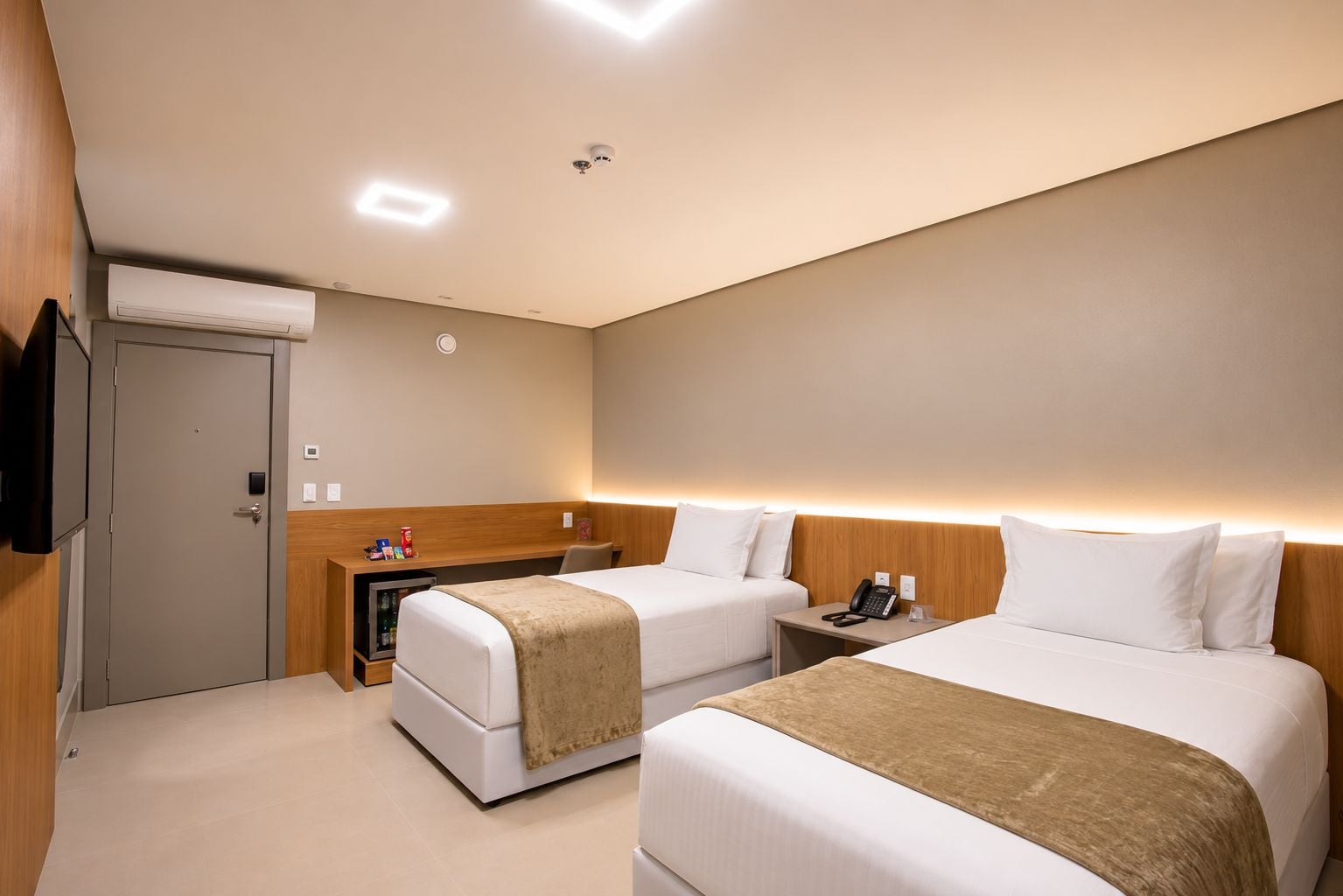 A hotel room with two beds, a bedside table with a phone, a small desk with a computer, and an air conditioning unit.