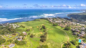 Vue aérienne d’un terrain de golf avec un paysage côtier en arrière-plan, mettant en valeur des verts et des allées bien entretenus, ainsi qu’un quartier résidentiel avec des maisons et une piscine.