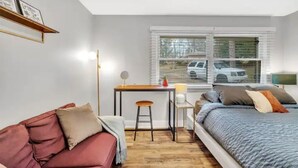 Un dormitorio pequeño con una cama, un escritorio y una silla. Tiene una ventana con persianas, un sofá rojo y una mesita de noche con una lámpara.