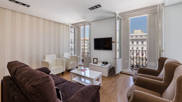 Un soggiorno con divano, poltrone, tavolino e televisore. È presente un balcone con ringhiera e vista sui palazzi.