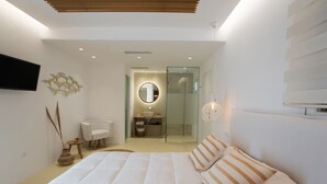 A modern bedroom with a large bed, a bedside table, a TV mounted on the wall, and a bathroom visible through a glass door. - Elit Suites Mykonos (Mykonos)