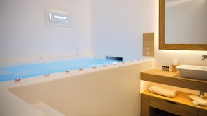A modern bathroom with a bathtub, a wooden vanity, a large mirror, and a wall-mounted light fixture. - Elit Suites Mykonos (Mykonos)