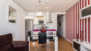 Een moderne woonkamer met een bruine bank, een glazen eettafel met zwarte stoelen, en een keuken met witte kasten, een rood aanrecht en roestvrijstalen apparatuur.