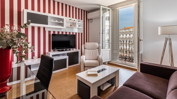 Een moderne woonkamer met een televisie, zitplaatsen, een boekenkast en een balkon met uitzicht op de stad.