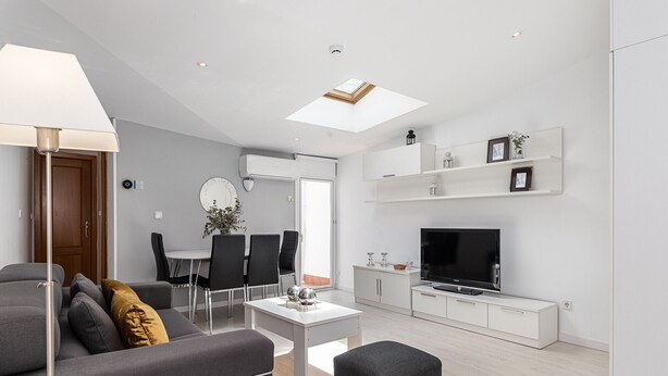 Una sala de estar moderna con sofá, mesa de centro y reposapiés. Hay un comedor con sillas y una mesa, y un televisor sobre un mueble blanco.