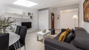 Una sala moderna con un televisor de pantalla plana, un comedor con mesa y sillas, un sofá con cojines, y un ventanal con vistas al exterior.