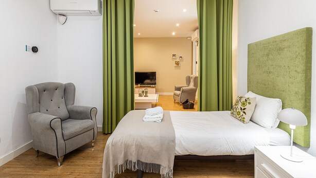 Un dormitorio con una cama, un sillón gris, un televisor y una cortina verde.