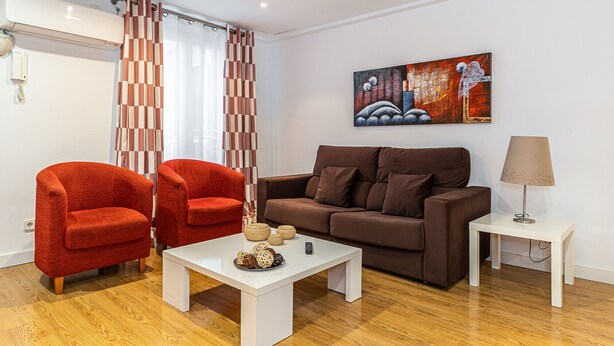 A living room with a brown sofa, two red armchairs, a white coffee table with decorative items, a painting on the wall, and a lamp on a side table. - Waou Apartamentos en Sol Confort 6 (Madrid)